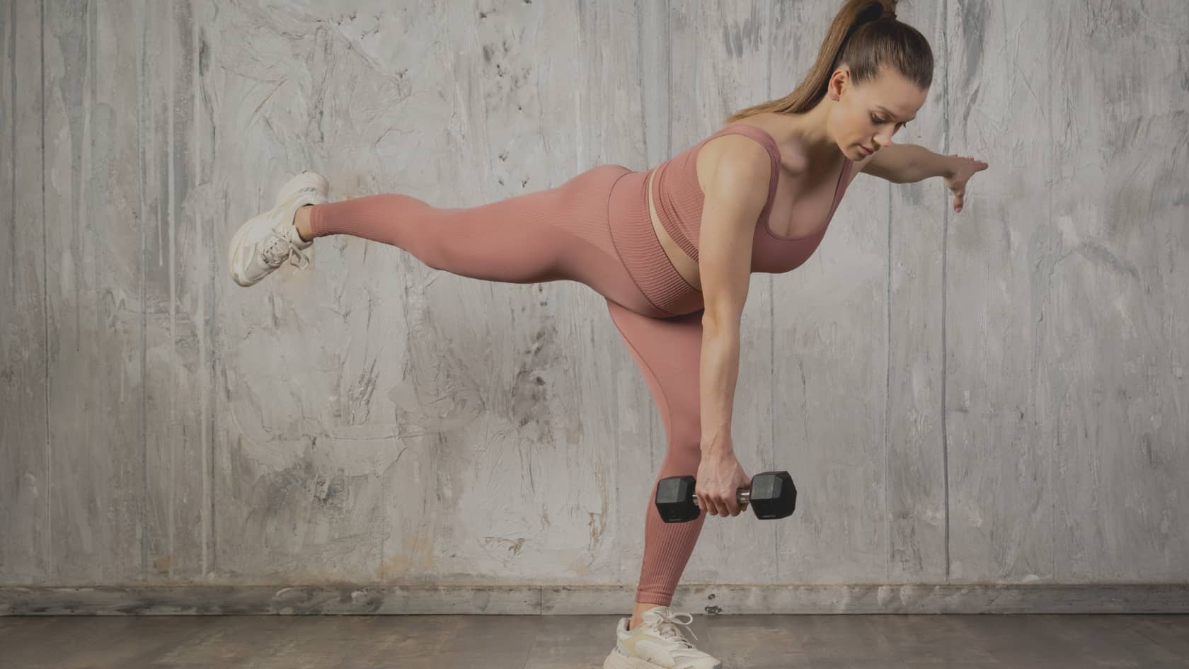 Une femme en tenue de sport seamless rose effectue un soulevé de terre sur une jambe (single-leg deadlift RDL) en salle de sport, le buste parallèle au sol avec un haltère noir à la main. Exemple d'exercice de renforcement applicable dans le cadre du sport prénatal adapté.