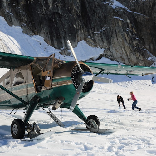 一架裝有滑雪板的綠色小型飛機停在雪地上，靠近一座岩石山；兩個人正在附近走動。