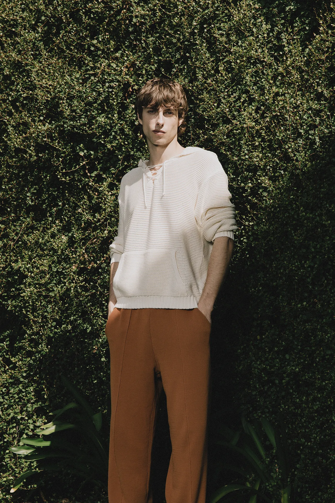 Annapurna SS26 collection men's cream white textured knit hoodie with lace-up neck and rust-colored trousers.