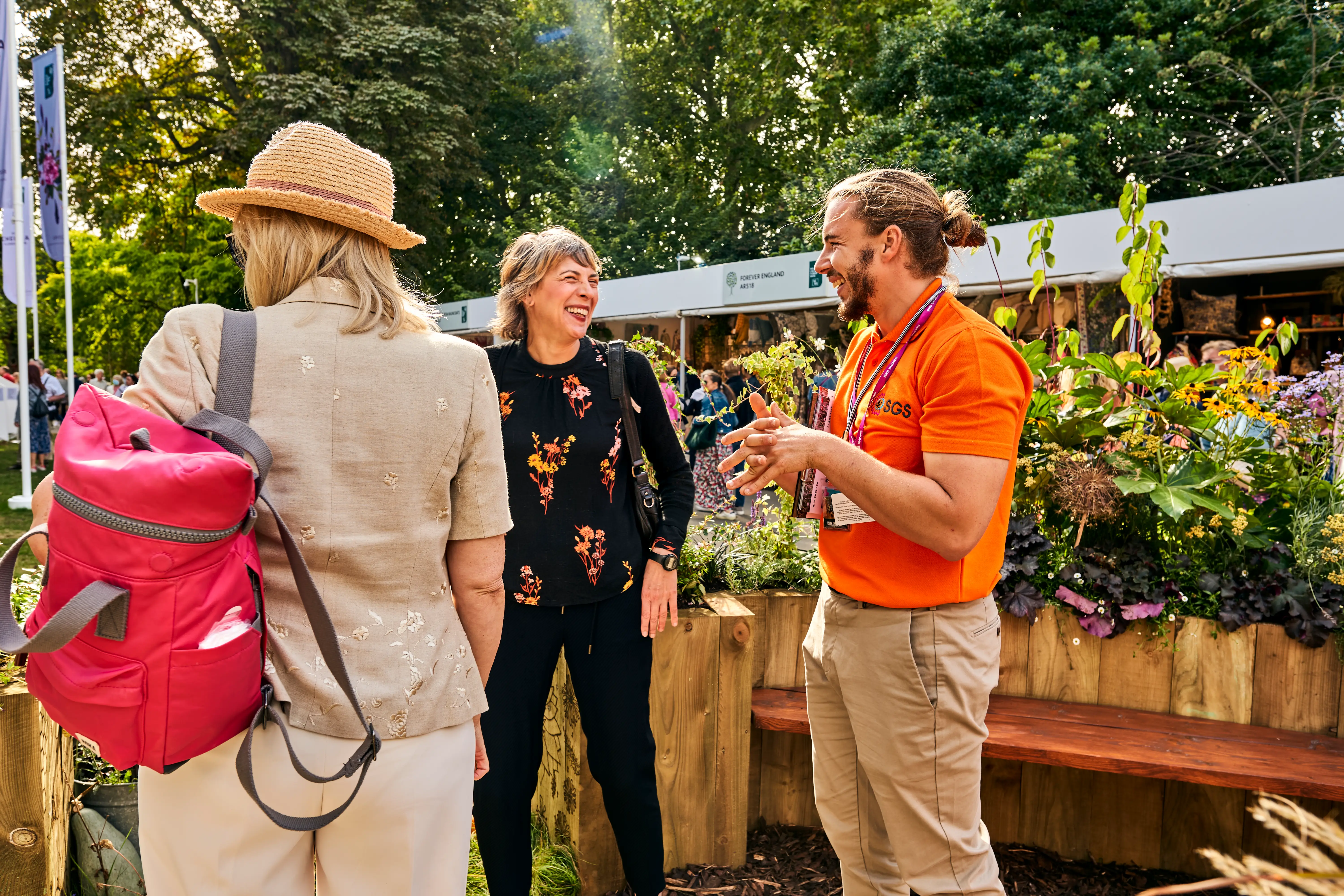 Three people engage in conversation outdoors, surrounded by greenery and a lively atmosphere.