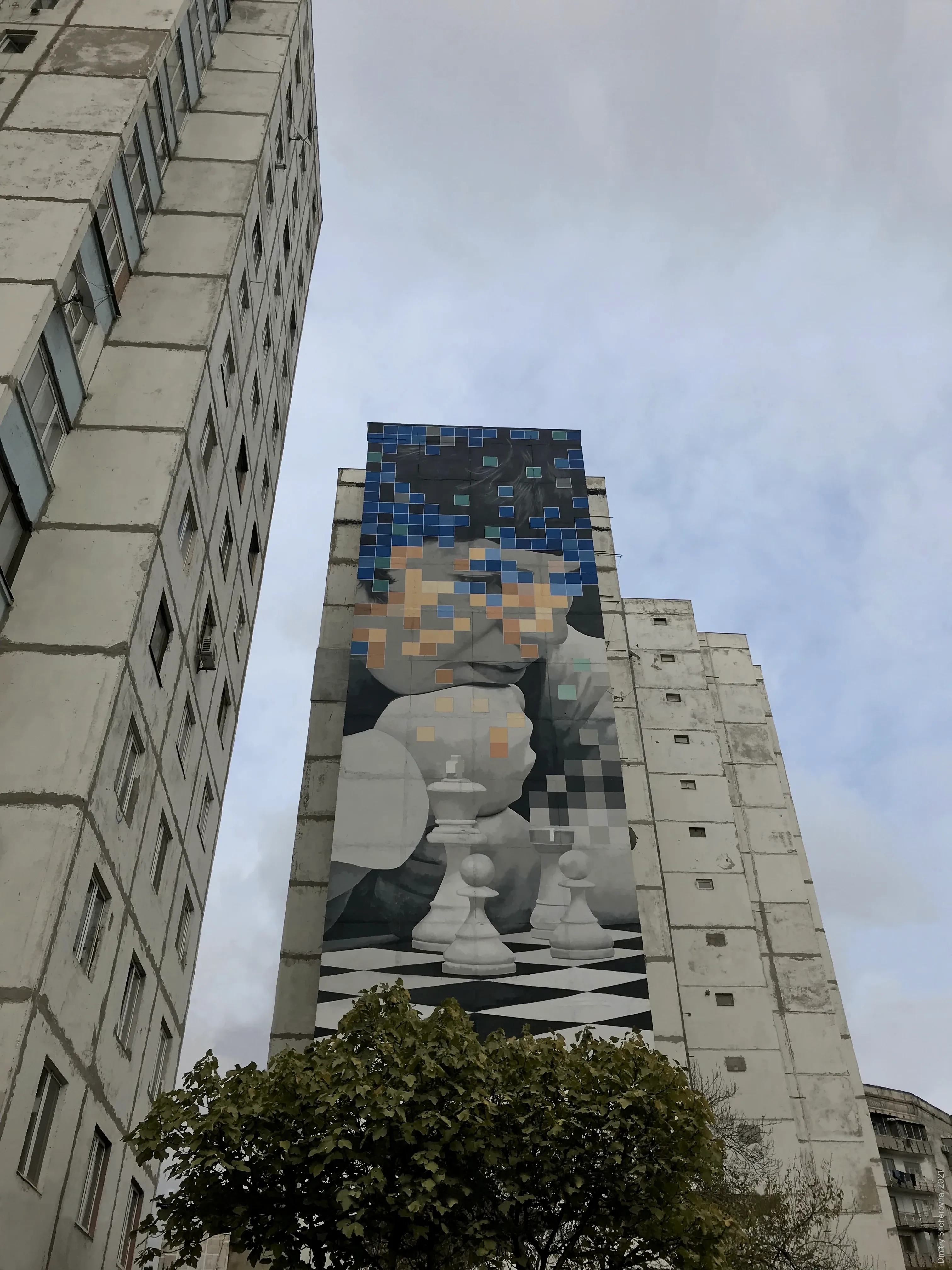 A tall mural on the side of a high-rise building depicts a figure leaning over a chessboard with white chess pieces. The figure's face dissolves into blue, teal, and tan pixel squares, blending realism with digital abstraction.