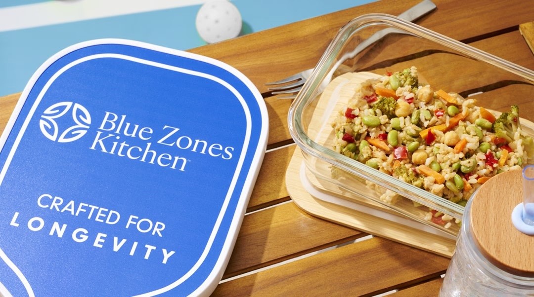Blue Zones Kitchen container and a glass dish of prepared grain and vegetable meal on a wooden table.