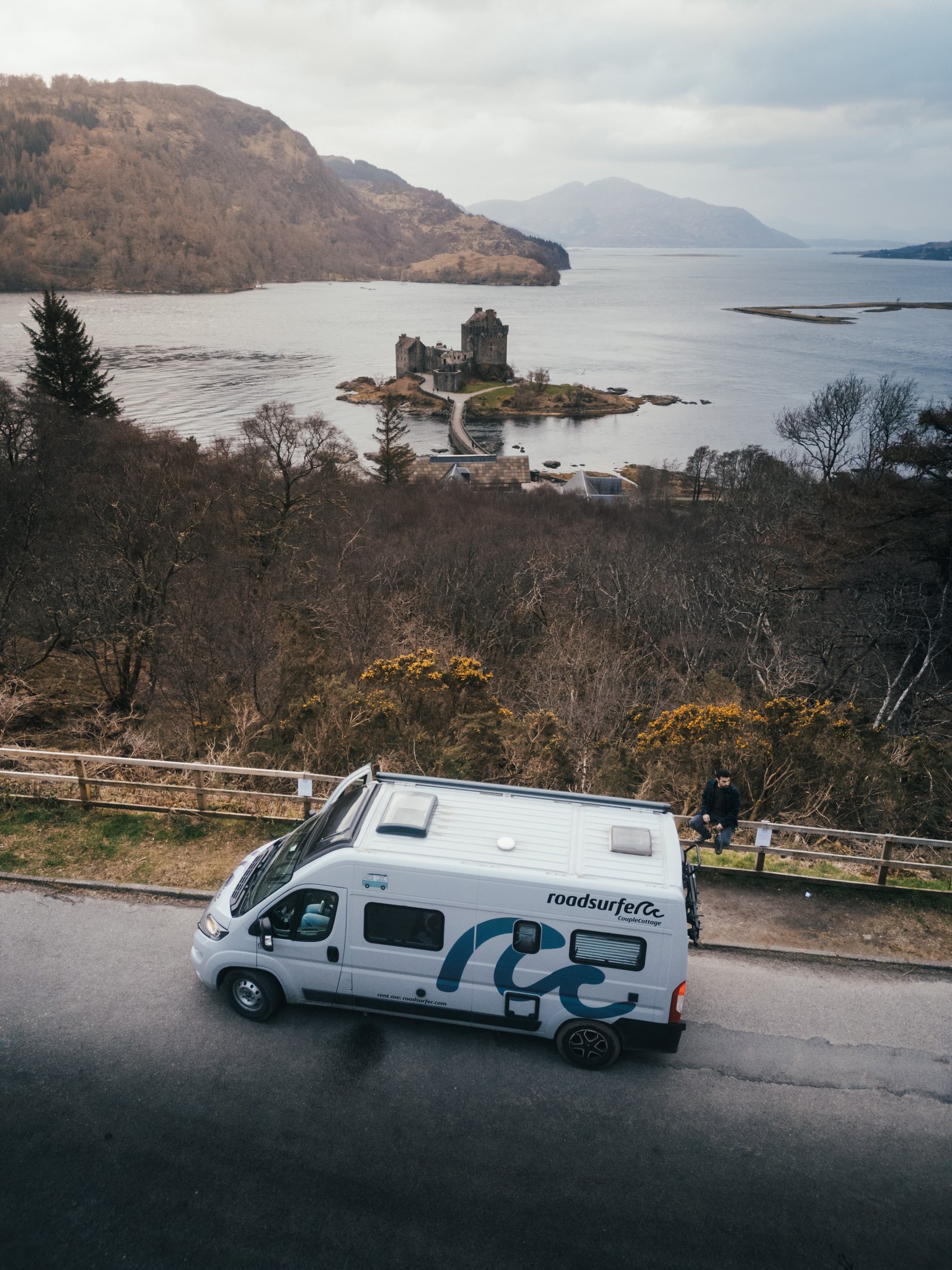 A parked camper van overlooks a scenic landscape with mountains and a body of water in the background.