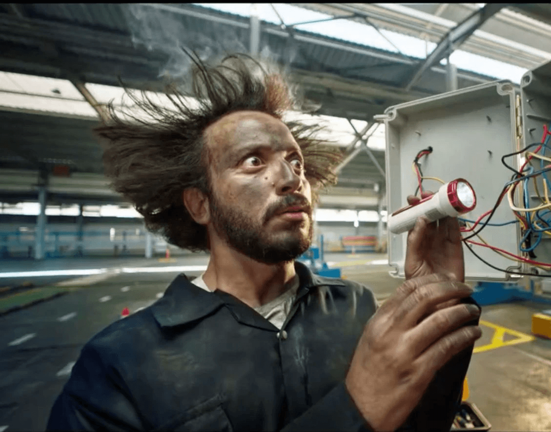 A surprised electrician with wild hair examines a flashlight and electrical box, smoke in the background.