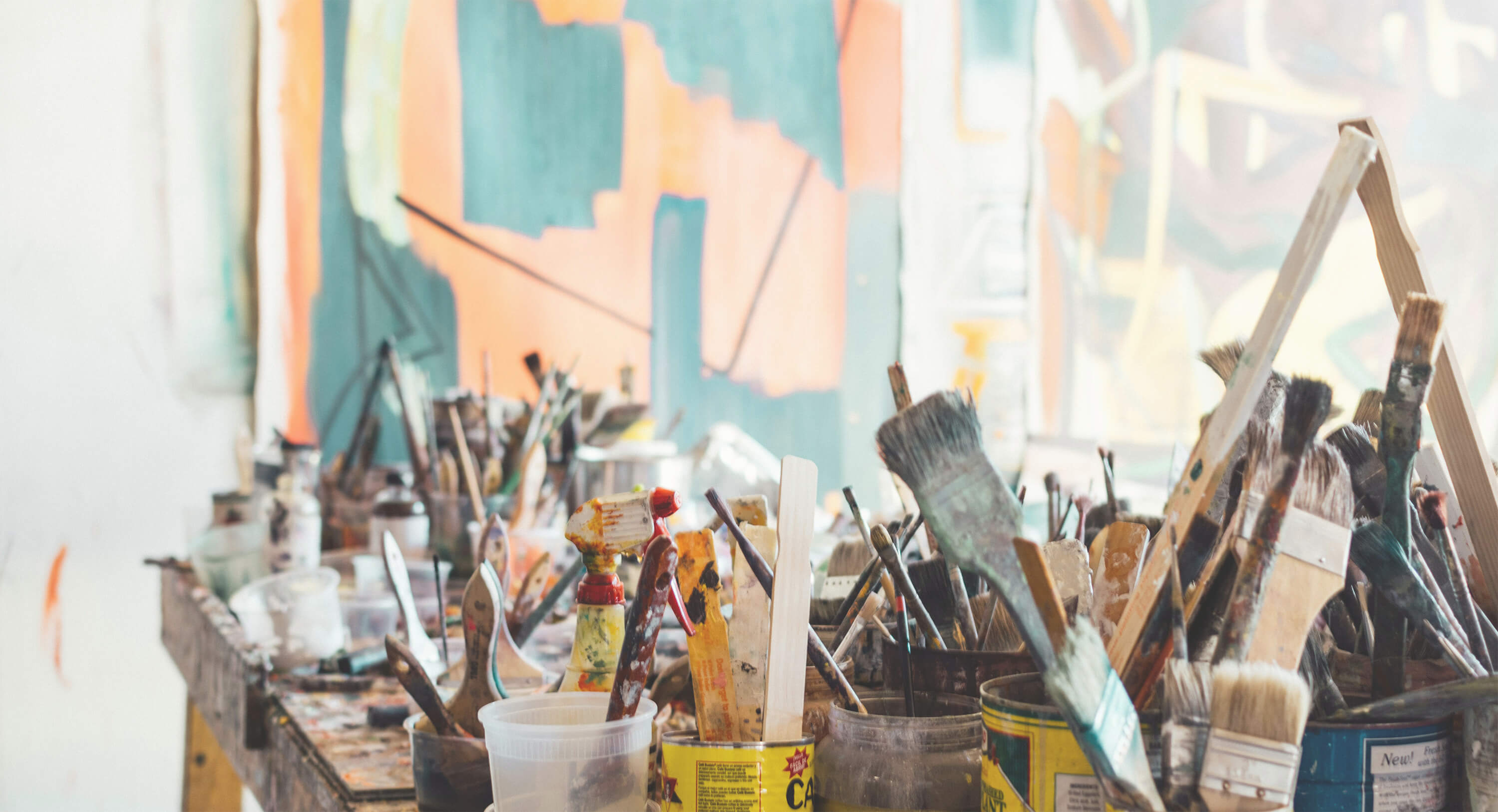 Artist's workspace with paint brushes in jars and canvas with abstract painting in teal, peach, and cream tones.