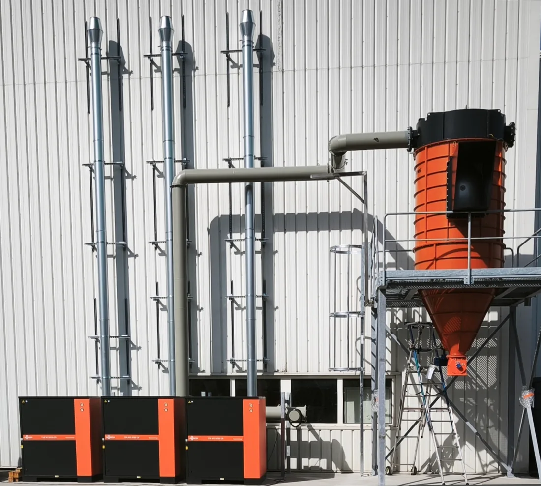Cyclone orange relié à plusieurs pompes et à des tuyauteries verticales pour l’aspiration industrielle en façade d’atelier.