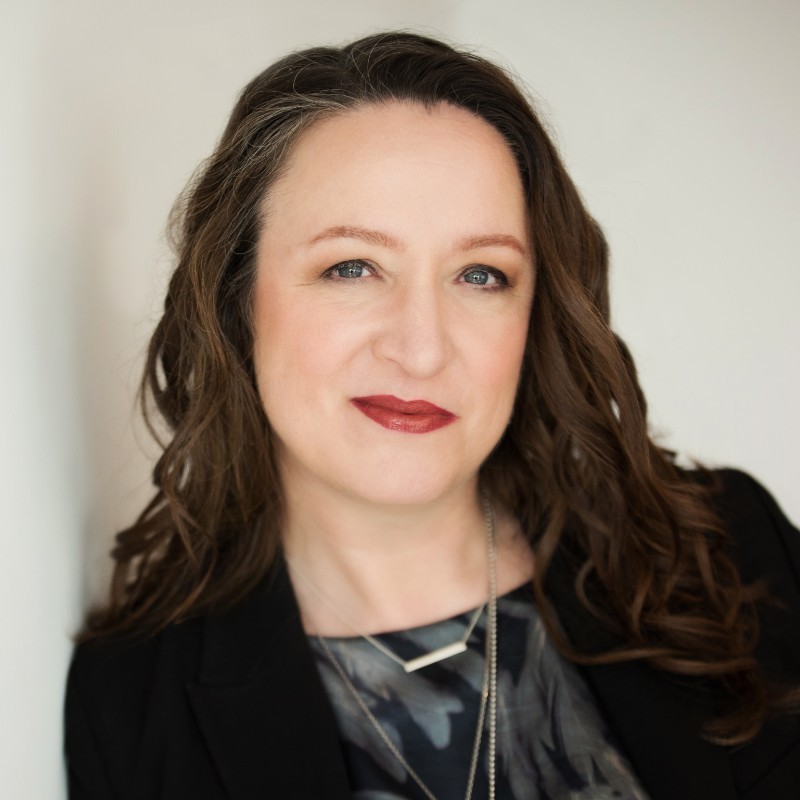 Smiling young man with curly hair, wearing a dark hoodie, against a softly lit background.