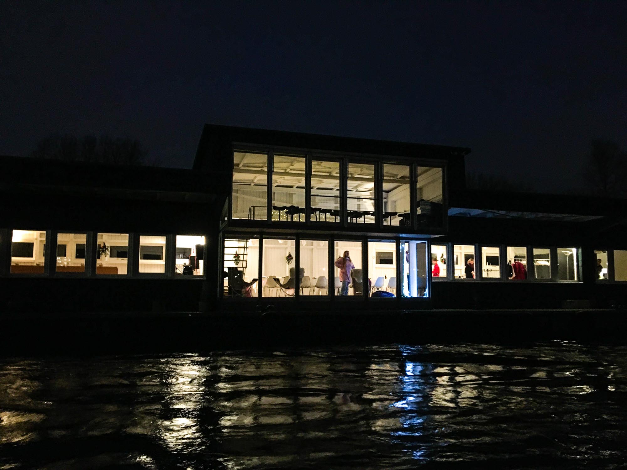 Blick von der Elbe auf den warm beleuchteten Ponton von Design for Human Nature in Hamburg auf dem Fluss.