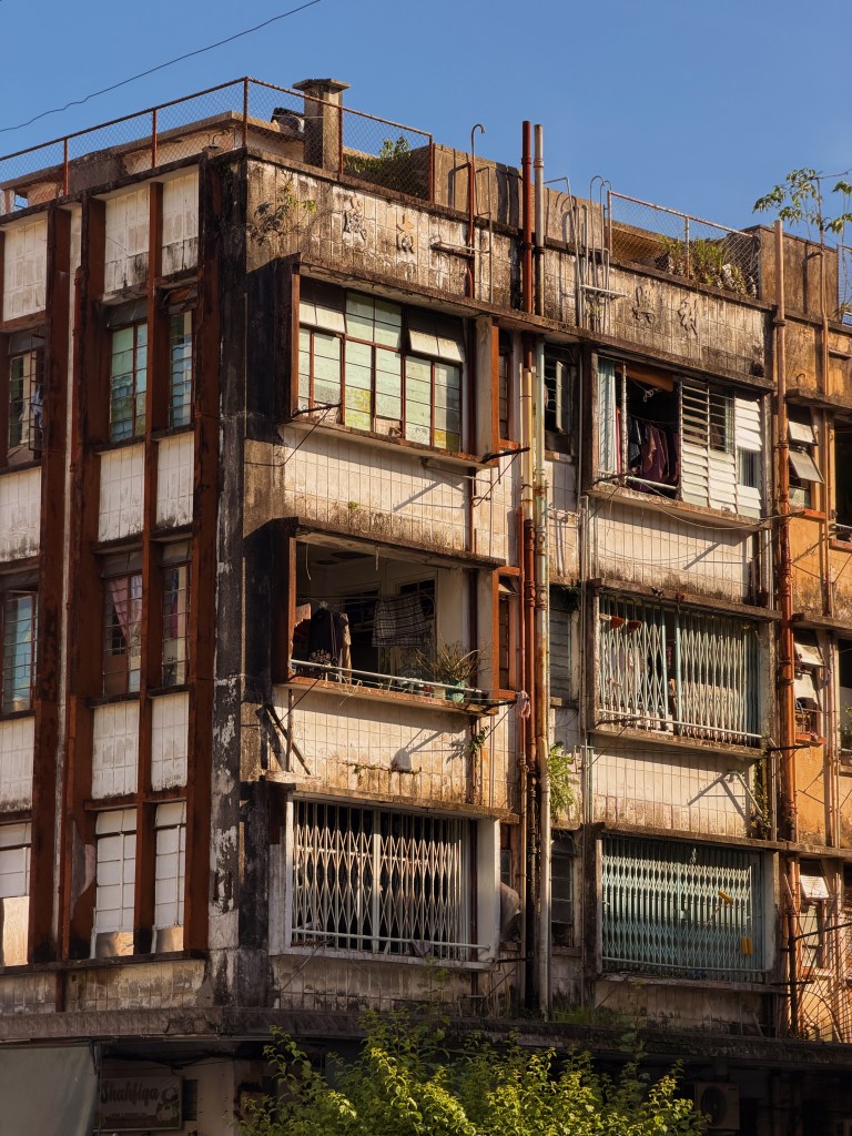 Close-up of a weathered building facade with a mix of rusted metal and peeling paint, featuring multiple windows and balconies adorned with plants.