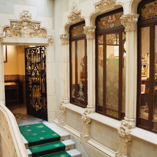 Ornate hallway with decorative arches, stained glass windows, green carpeted stairs, and an intricate iron gate.