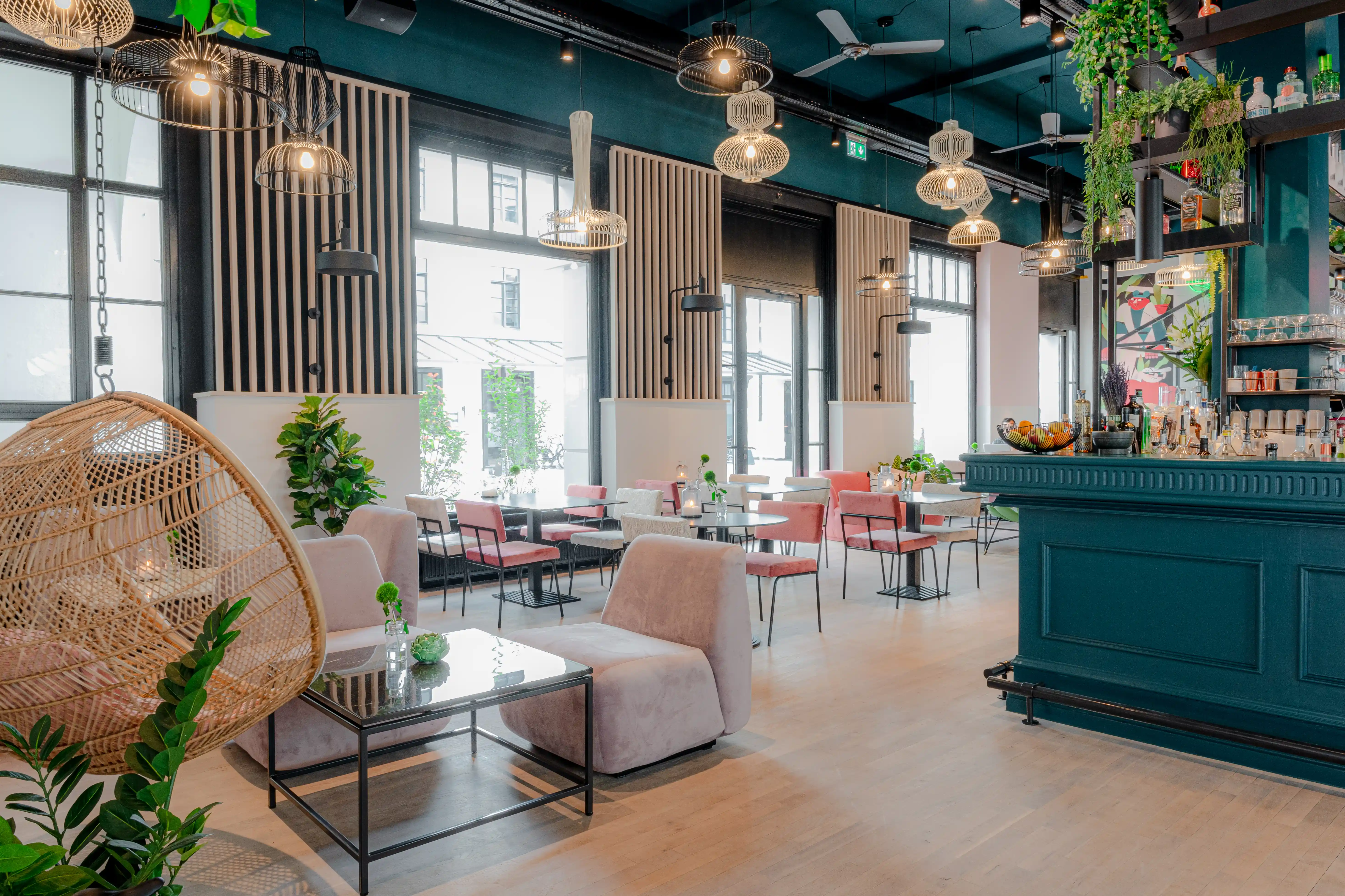 Overview of LOKAL VIER with a hanging rattan egg chair, pink lounge chairs and glass tables, slatted wall panels, woven pendant lamps and the teal bar on the right.
