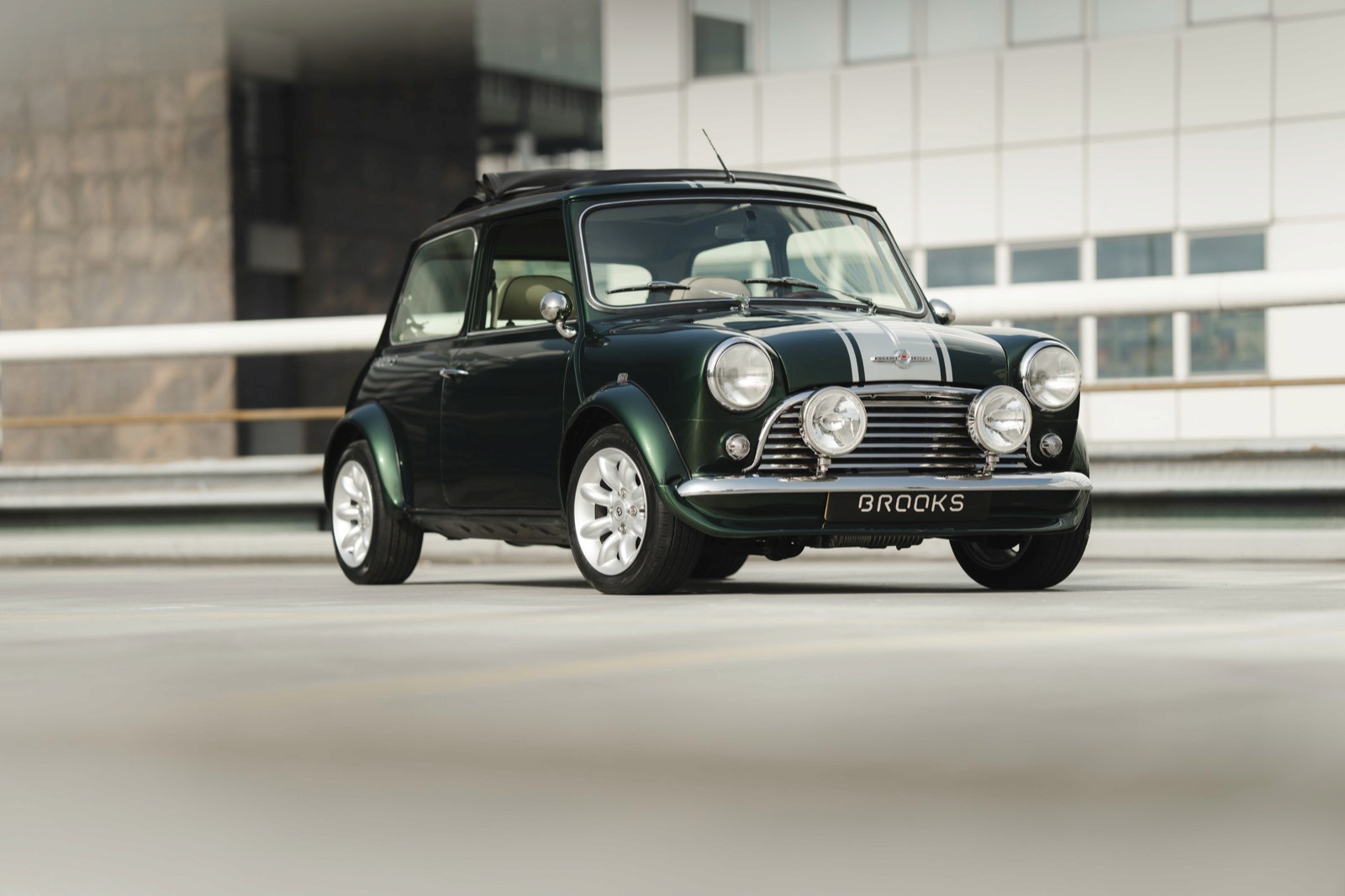 A vintage green car driving on a road with a modern building in the background.