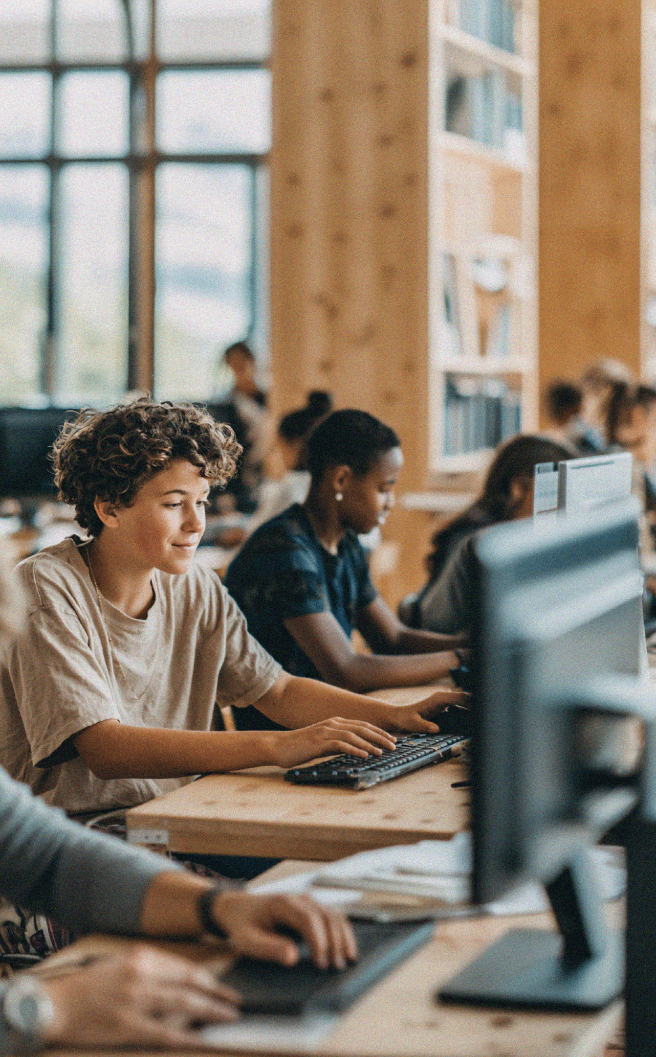 Several young people sit at desktop computers in a bright classroom or computer lab. In the foreground, one student with short, curly hair types on a keyboard while looking at the monitor with a slight smile. Other students work at nearby stations, and large windows and wooden walls create a warm, modern learning environment.