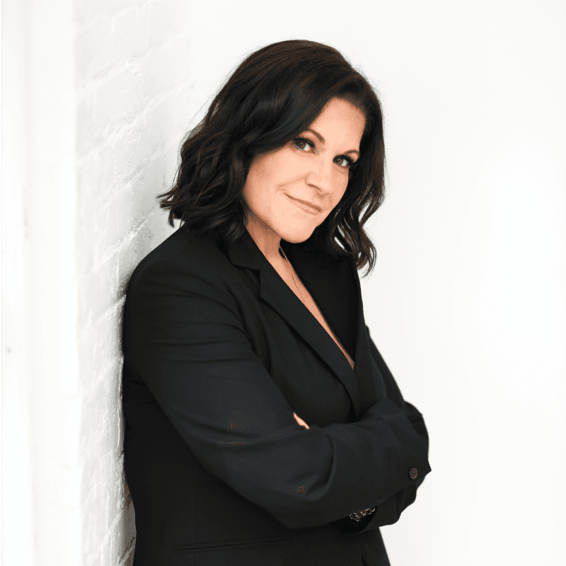 A headshot of a white woman with short black hair, smiling, wearing a black suit coat and leaning up against a white wall with her arms crossed