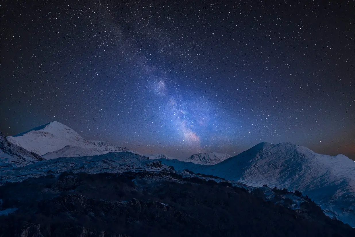 A starry night sky with the Milky Way over dark mountains and snow-covered peaks.