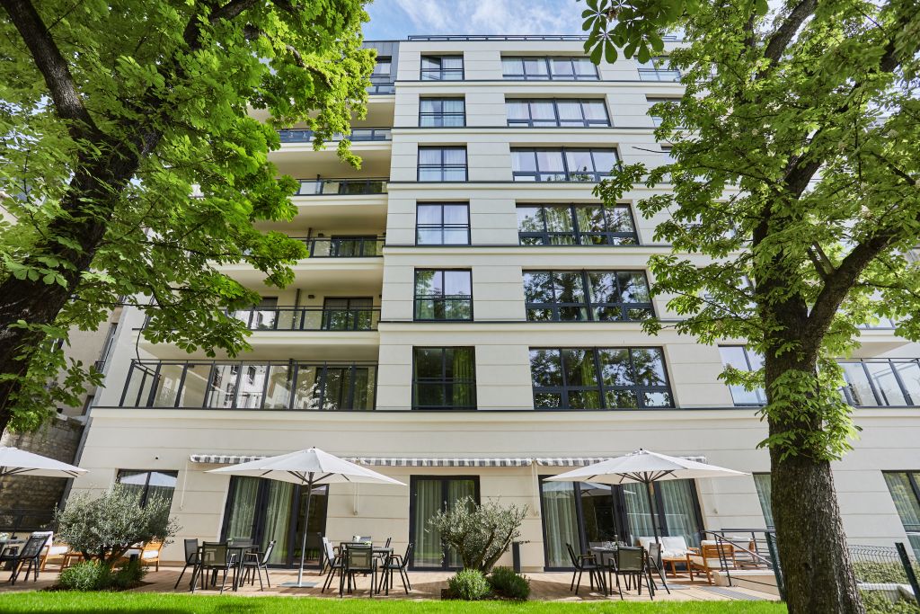 Modern multi-story residential building with green ivy covering left side, white balconies, large windows, and lush vegetation in foreground