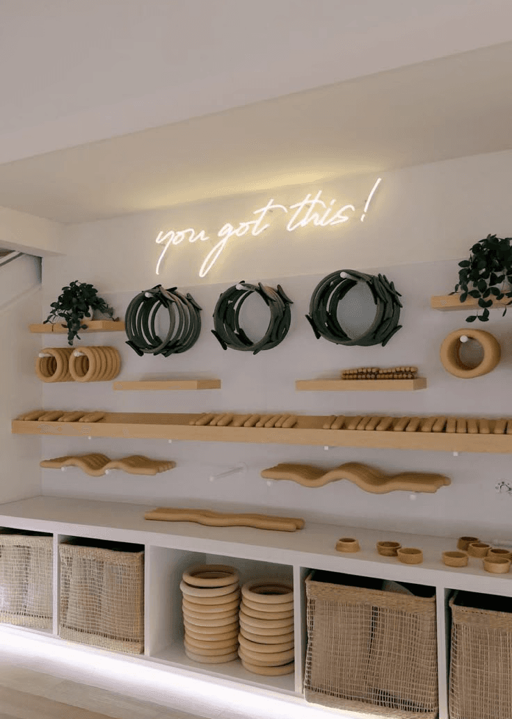 White cursive neon sign reading “you got this!” mounted on a Pilates studio wall above neatly arranged reformer rings, wooden exercise tools, and woven storage baskets.