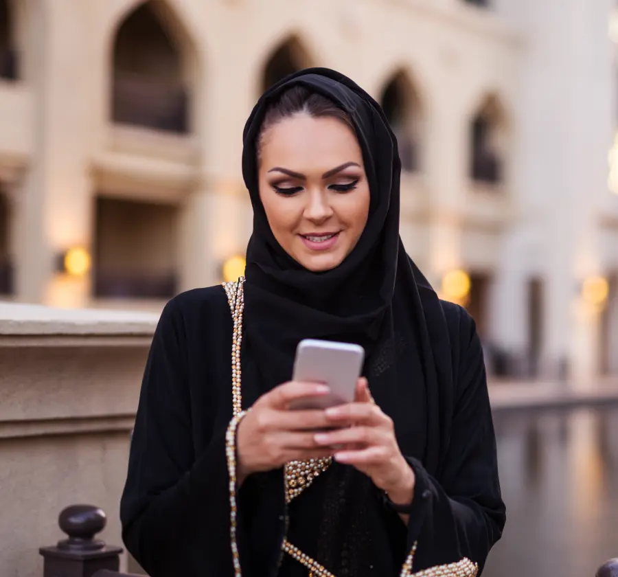 A woman in a black abaya smiling while using a health app on her smartphone outdoors, symbolizing the accessibility and convenience of digital health solutions for managing chronic conditions.