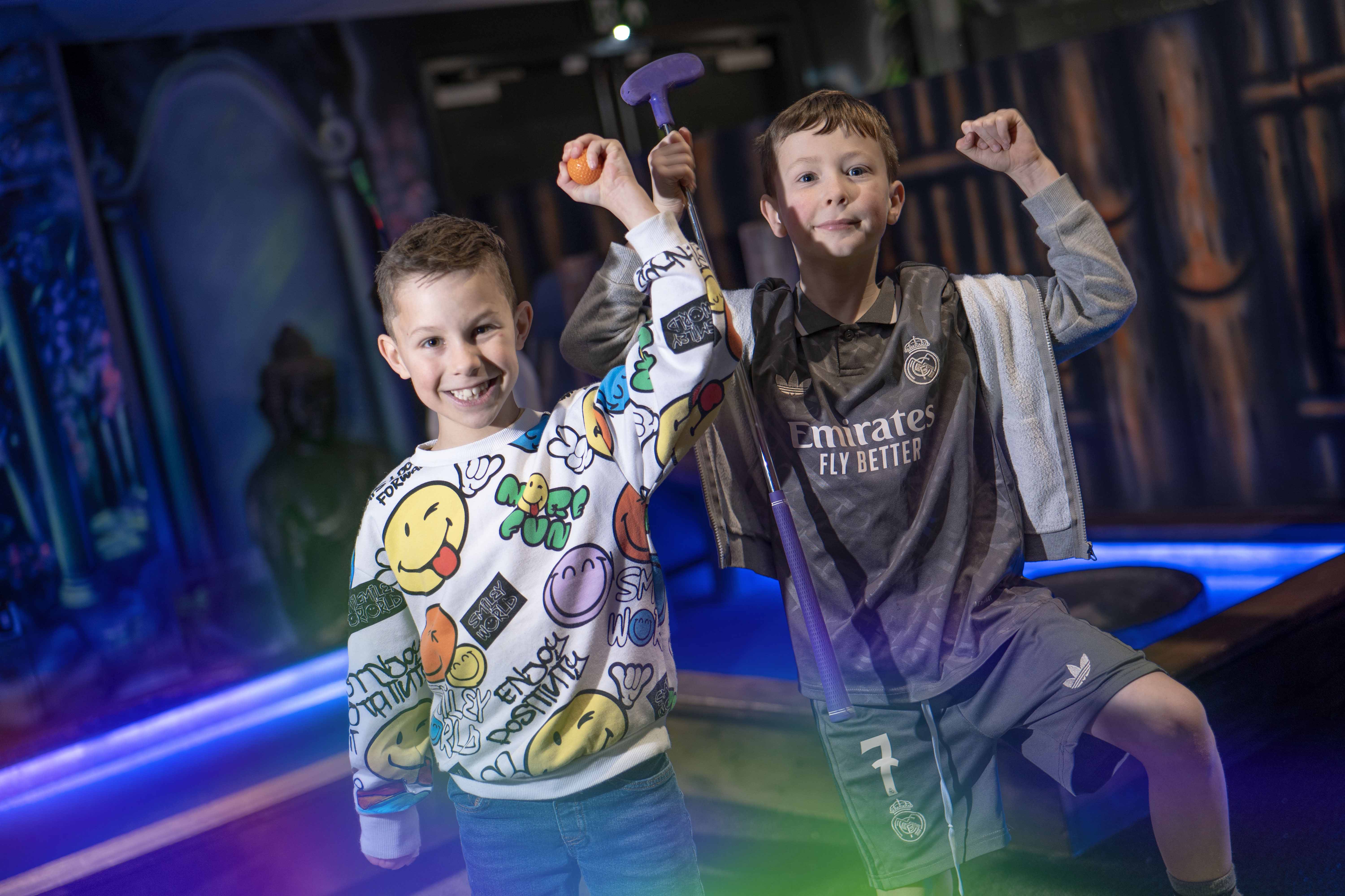 Two young boys cheerfully pose with colorful smiles in a vibrant, graffiti-adorned arcade setting, holding game tokens and prizes, embodying playful excitement and youthful energy.
