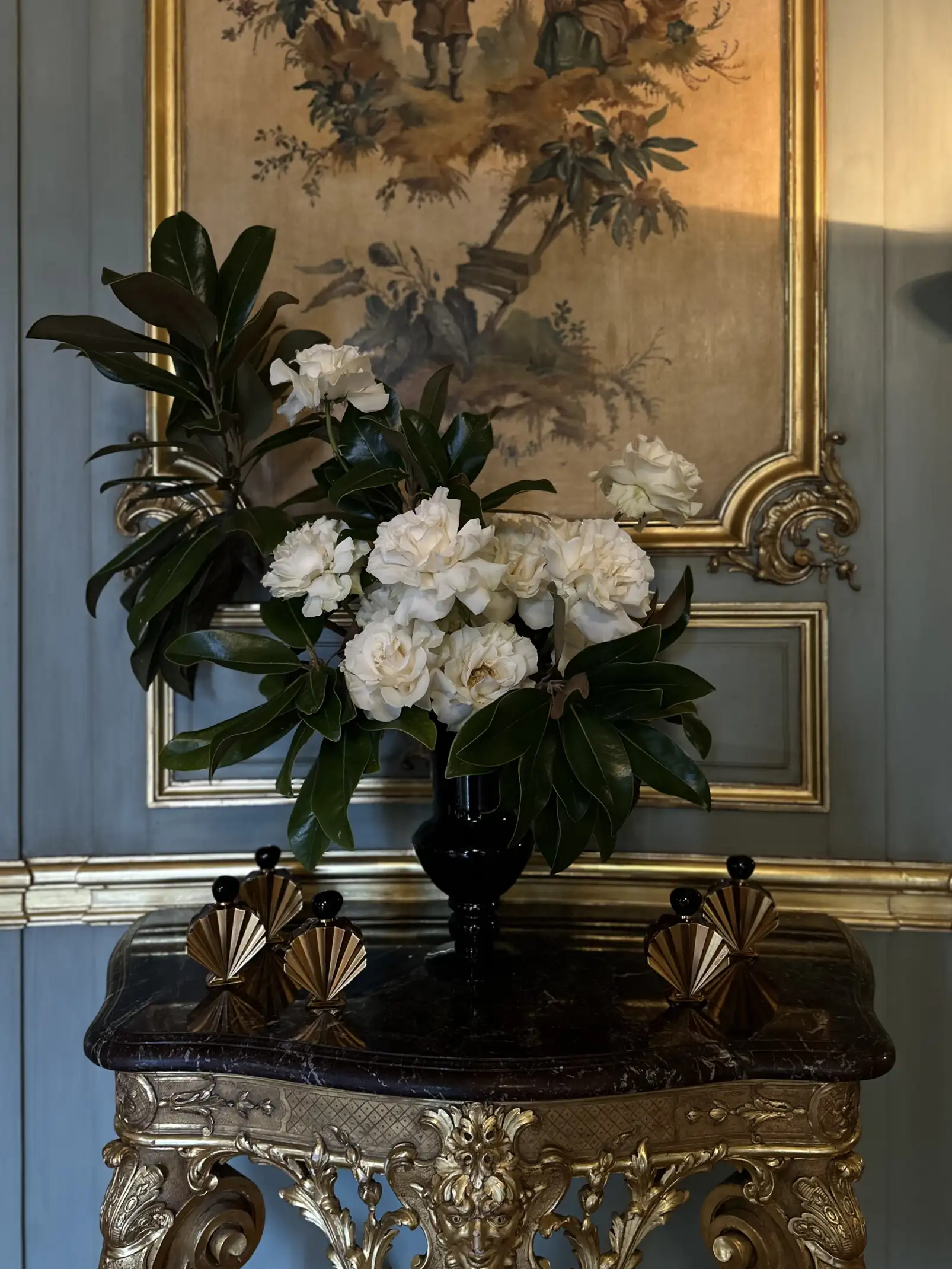 Arreglo floral con flores blancas y hojas verdes brillantes en un jarrón negro, colocado sobre una mesa de mármol oscuro con base dorada tallada, frente a una pared azul con molduras doradas y una pintura clásica enmarcada.