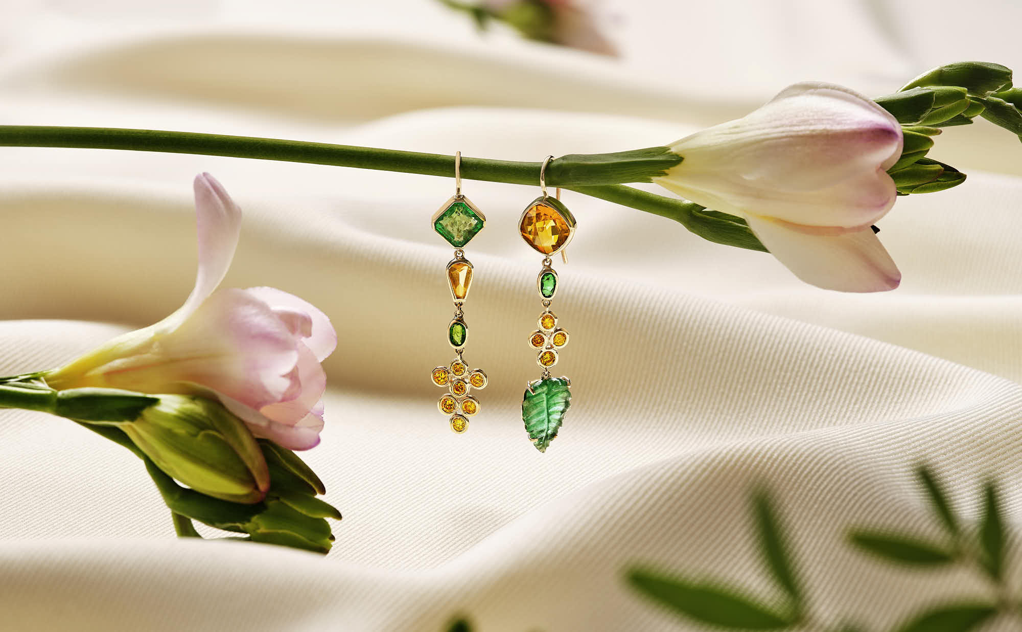 Colorful earrings displayed on soft white fabric, surrounded by delicate flowers and green leaves.