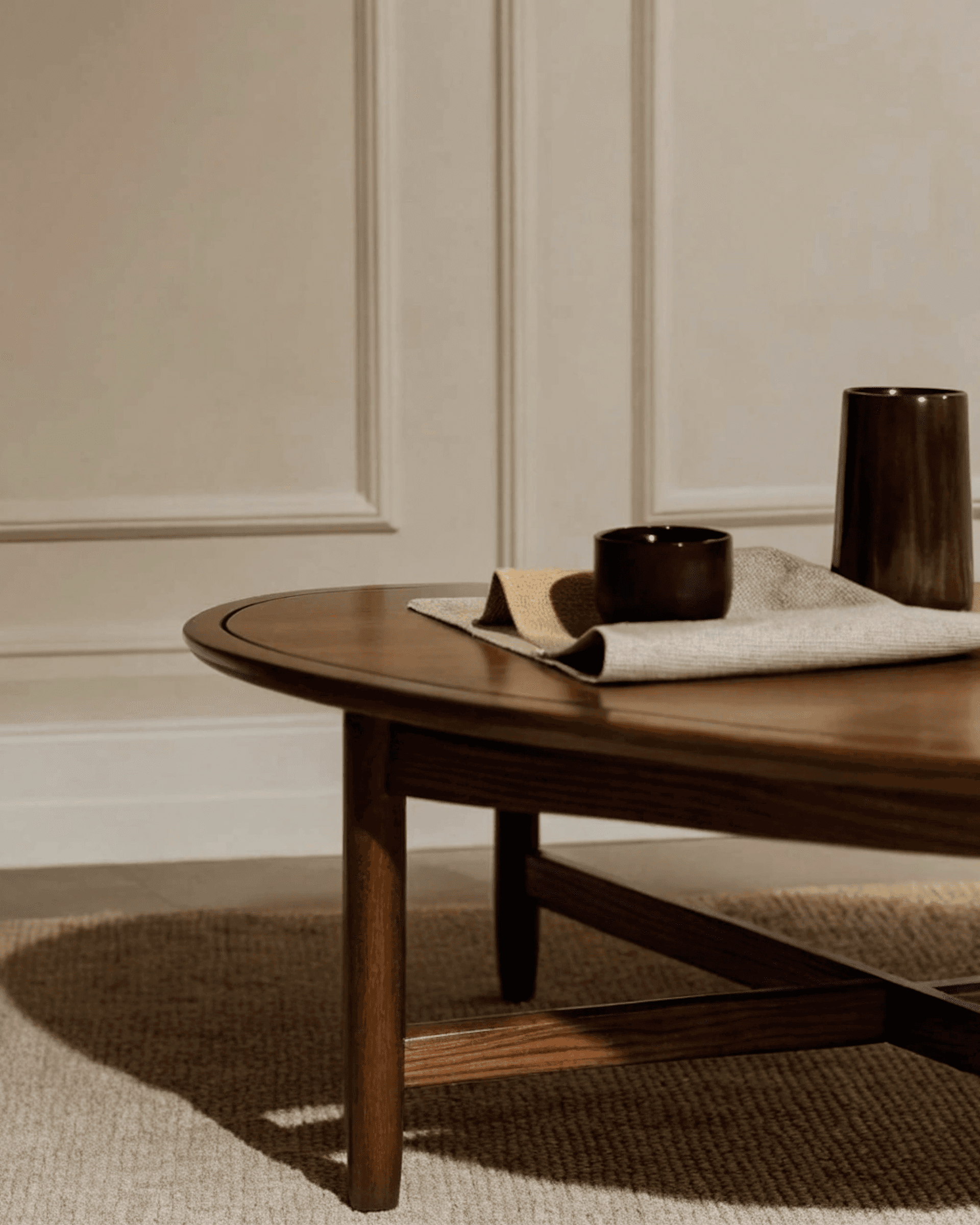 Minimalist wooden coffee table with dark ceramic vessels on a beige rug, set against a cream paneled wall. Warm, natural lighting.