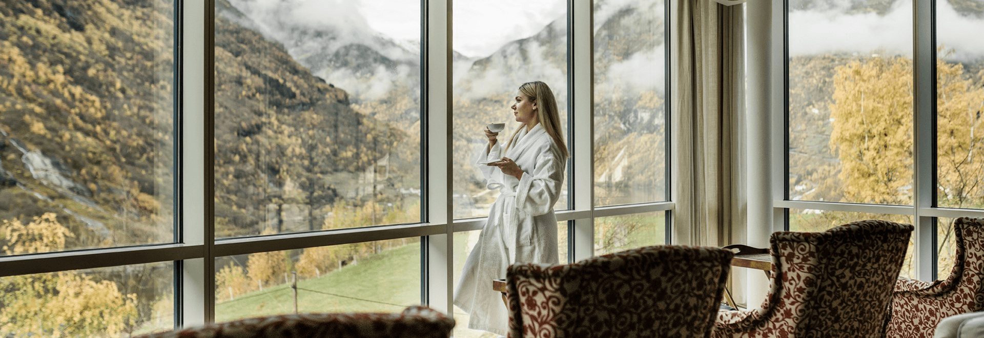A woman in a bright room gazes out large windows, overlooking a scenic mountain landscape.