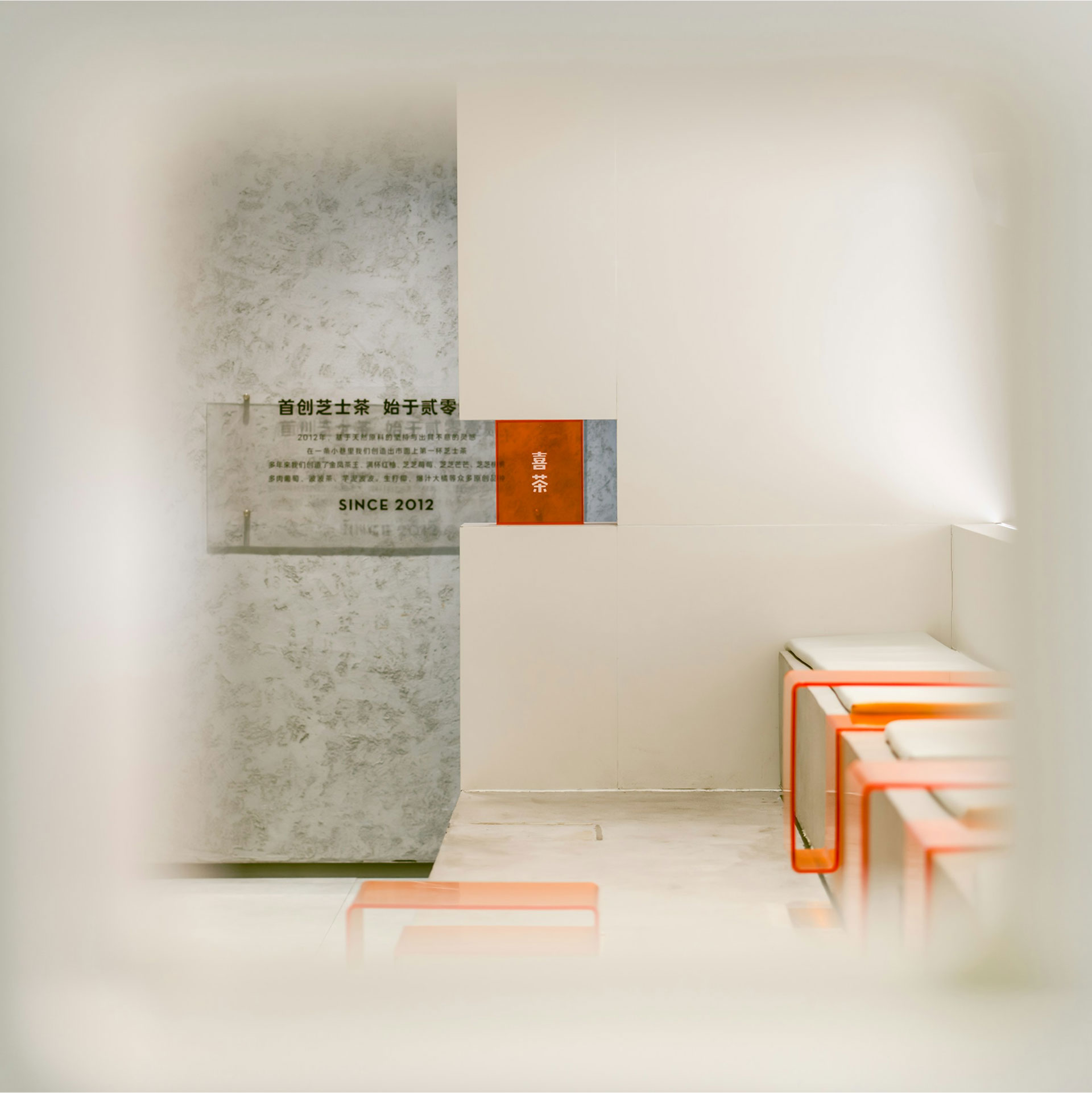 Minimalistic, modern interior design features soft white walls, geometric orange accents on block seating, and a smooth gray marble-like textured wall with a subtle plaque, creating a clean, contemporary atmosphere.