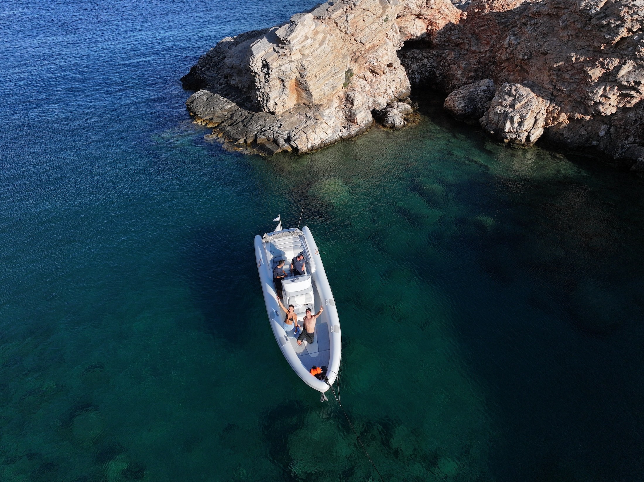 White motor yacht anchored in crystal-clear turquoise waters near dramatic rocky coastline, with guests relaxing on deck.