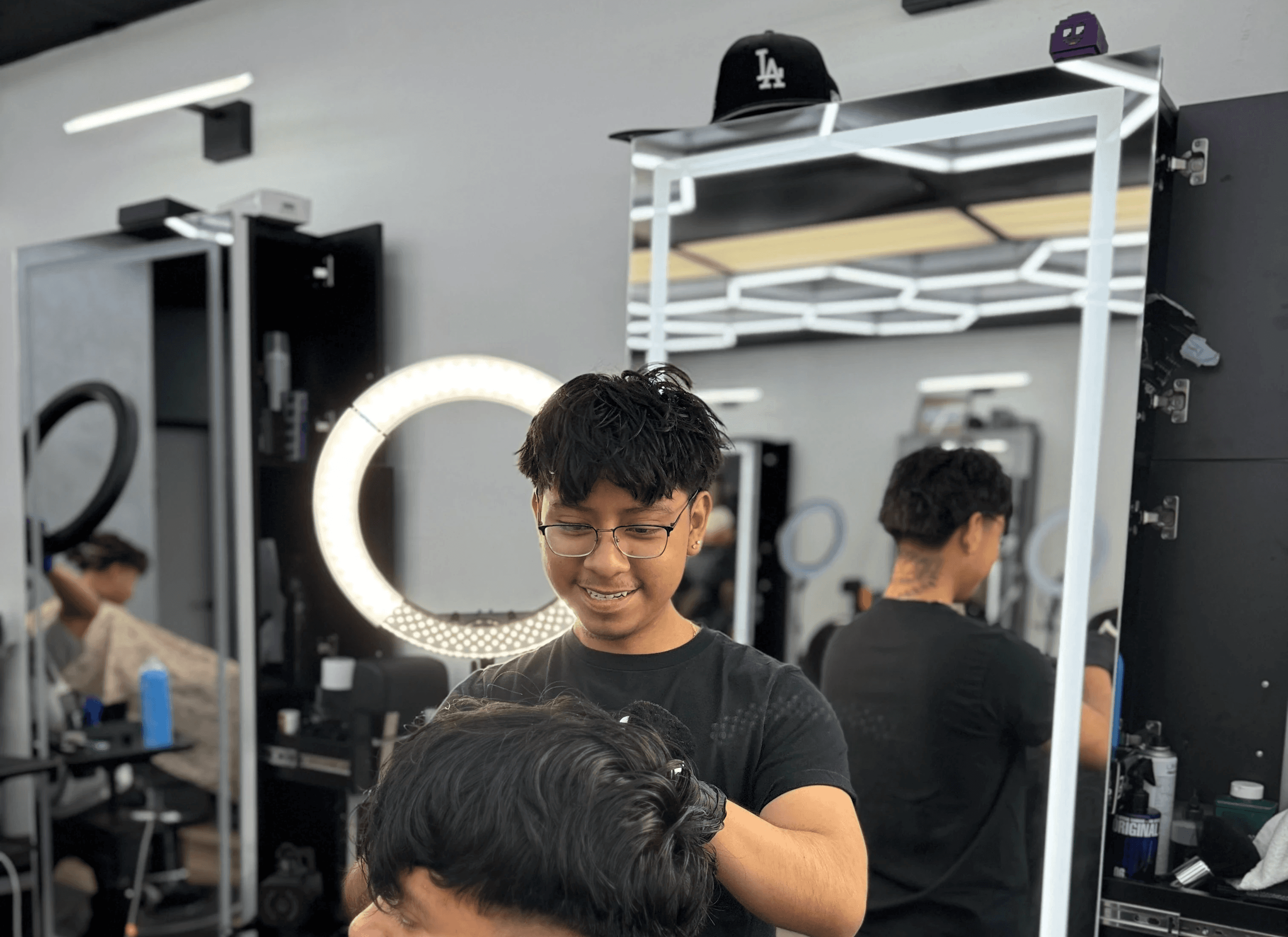 A barber shapes a client’s haircut using clippers near a bright ring light in barbershop. 