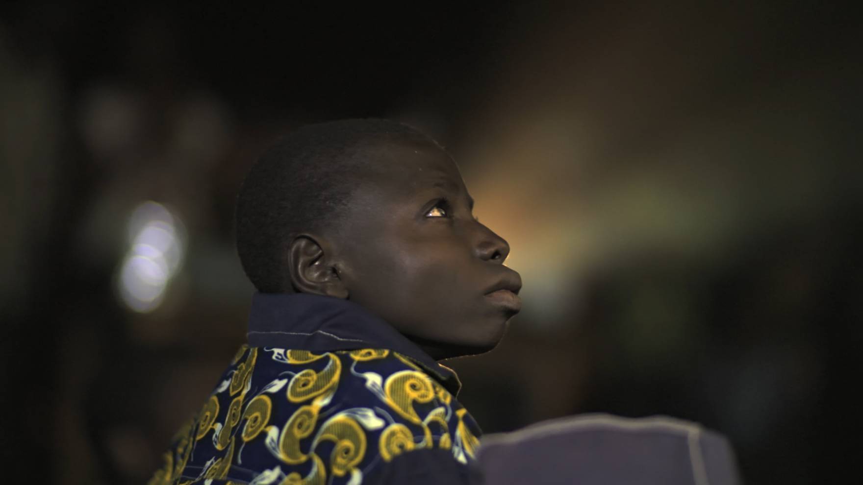 A young boy gazes upward with a thoughtful expression, wearing a dark shirt adorned with bright, swirling patterns. The background is softly blurred.