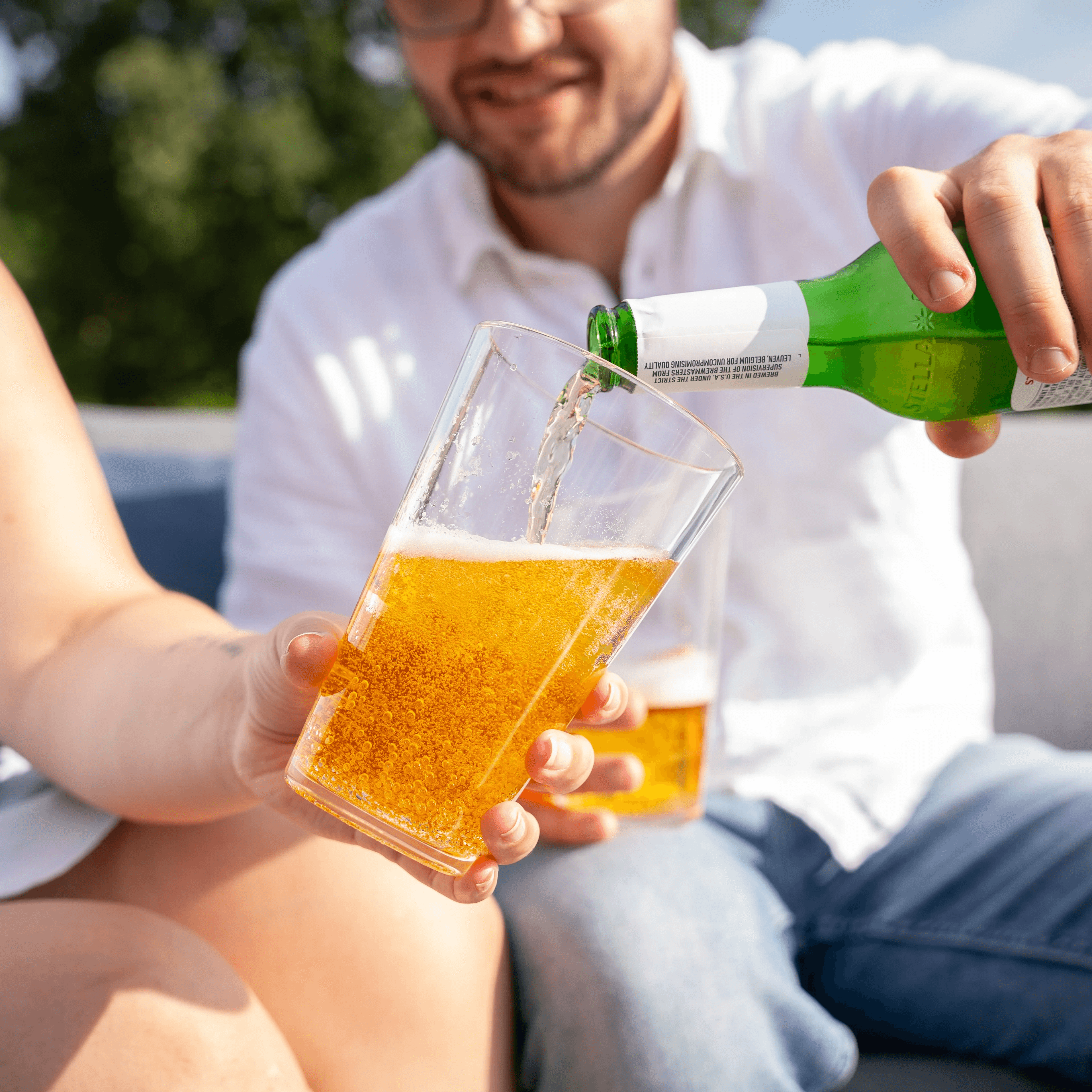 Two people in the image fram. The person closest to the camera lens is holding a translucent silipint pint cup that resembles glass while th eperson that is most in view but furthest from the camera is pooring beer into that cup.