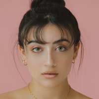 Portrait of a young woman with dark hair in a bun, wearing small gold hoop earrings and a delicate gold necklace, against a solid pink background.