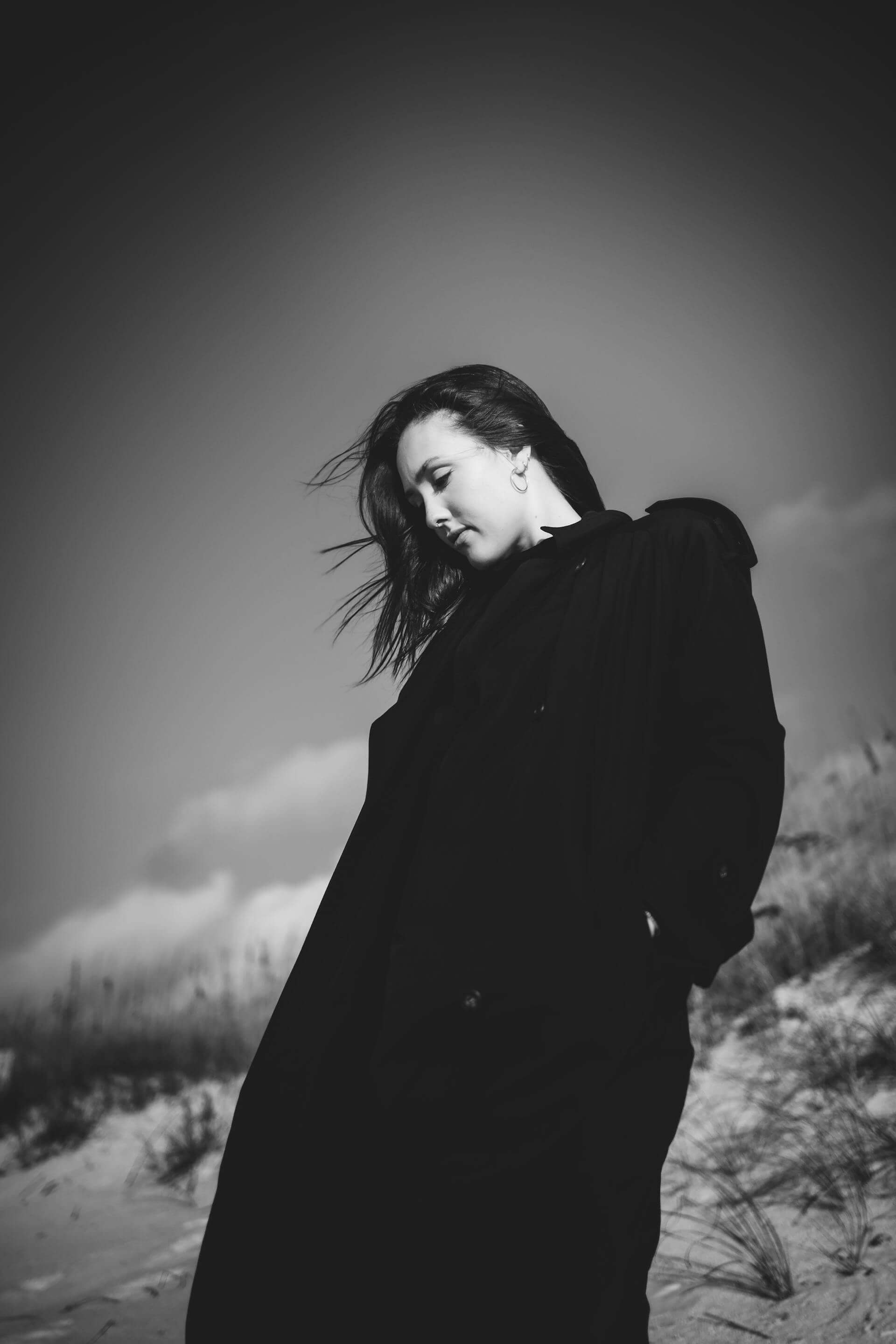 A woman in a black coat stands on the beach, gazing at the waves under a cloudy sky