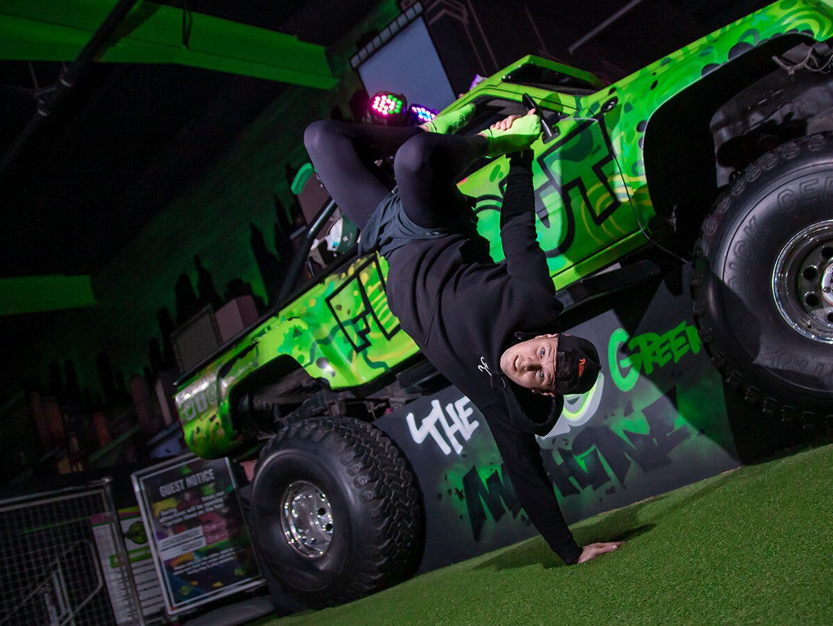 A breakdancer performs a handstand in front of a bright green monster truck. 