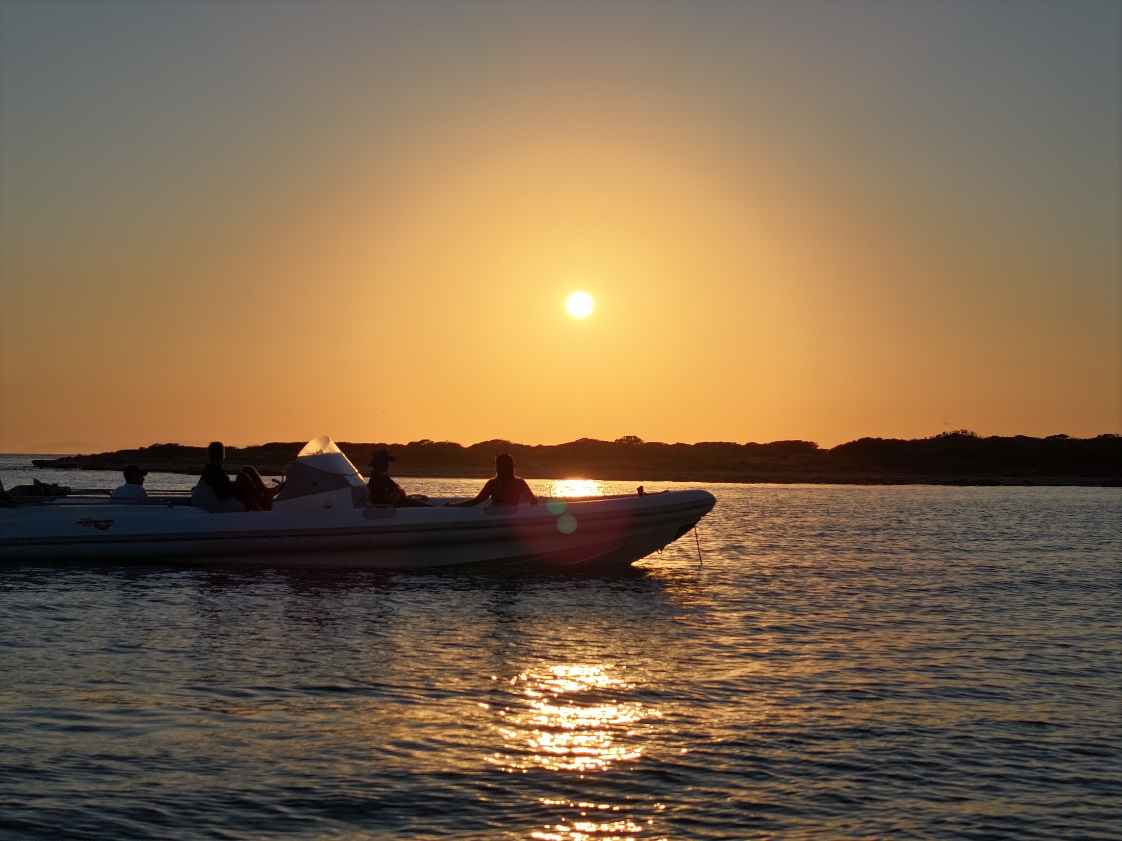 White speedboat with passengers cruising on golden waters during sunset, creating a shimmering reflection path across the calm sea.
