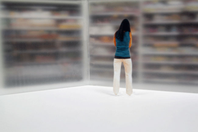 A miniature figurine of a woman with long dark hair, wearing a teal sweater and light-colored pants, stands on a white surface facing a large, out-of-focus bookshelf. The figurine is positioned with its back to the viewer, facing the bookshelf. The bookshelf appears to contain books or similar items, though their details are blurred due to the depth of field. The background is predominantly white, emphasizing the figurine and the bookshelf.