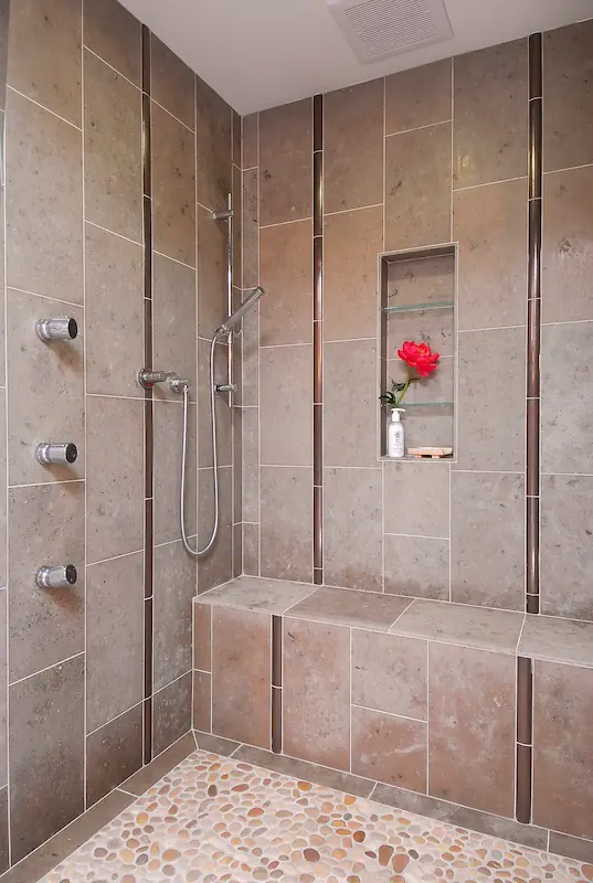 Primary bath walk-in shower enclosure with tile walls, niche and bench, and stainless steel fixtures.Modern kitchen with wooden cabinets, central island, hanging lights, and dark flooring. Irvine Transitional Modern. Photo by Chris Darnall.