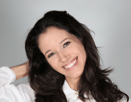 A woman with long, dark brown hair is captured smiling at the camera. She is wearing a white collared shirt and has her right hand raised near her head. The background is a plain light gray or off-white color. Her eyes are dark brown or black, and her teeth are visible in her smile.