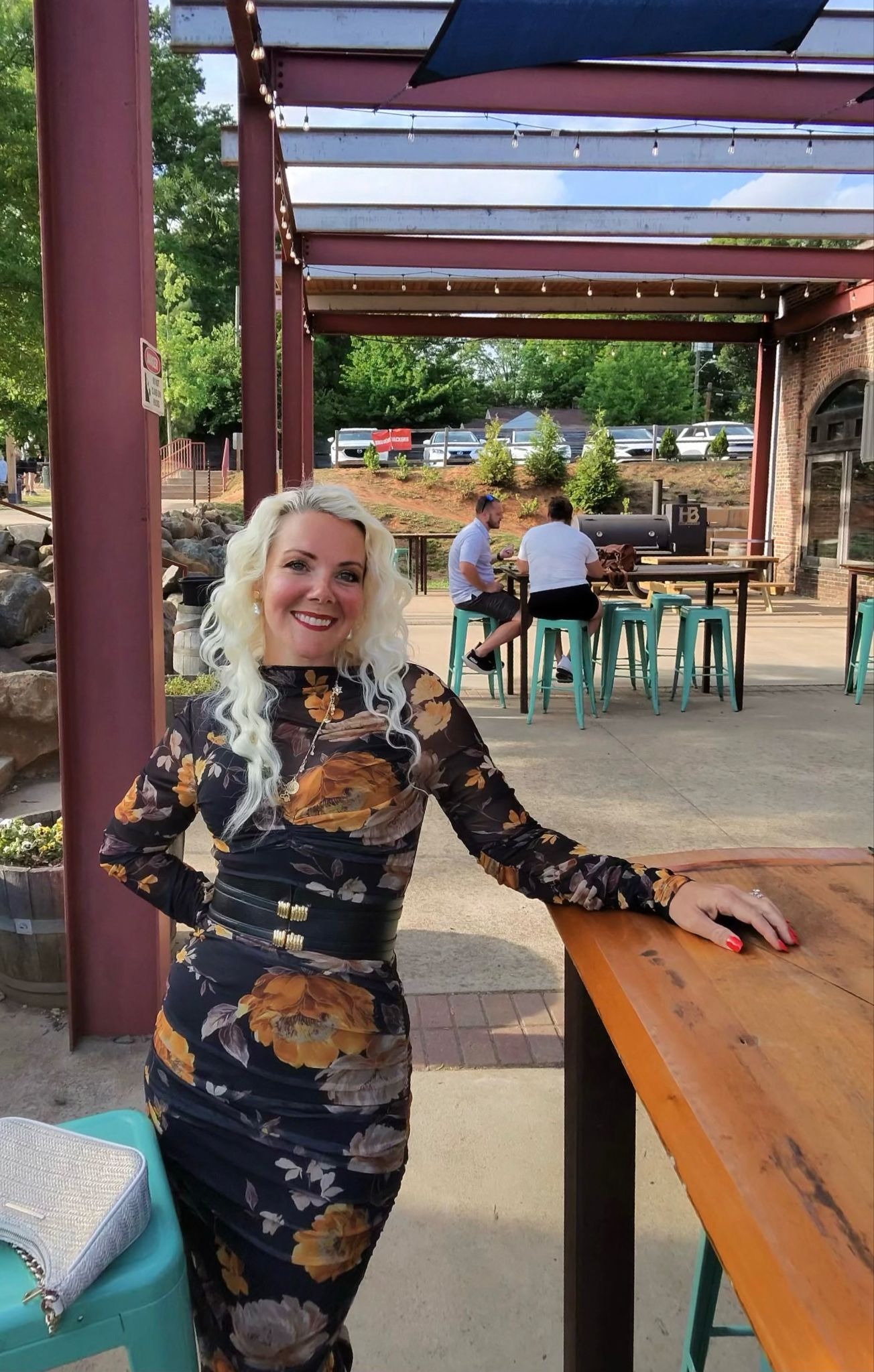 Smiling woman in floral dress standing at outdoor patio with tables and string lights.