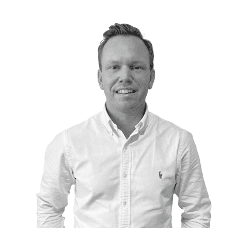 A focused woman with long red hair is seated at a desk, wearing a black blazer and dark blouse. She intently looks at camera, smiling slightly, with an open laptop in front of her. The setting is a warm and serene office setup in the background. Soft daylight filters through, creating a calm atmosphere. The composition captures her in a frontal profile with a clear emphasis on her concentration and the serene indoor setting.