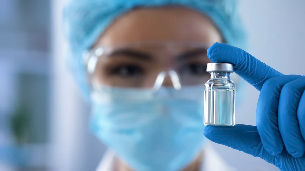 Scientist in a mask and safety glasses holding up a small clear glass vial.