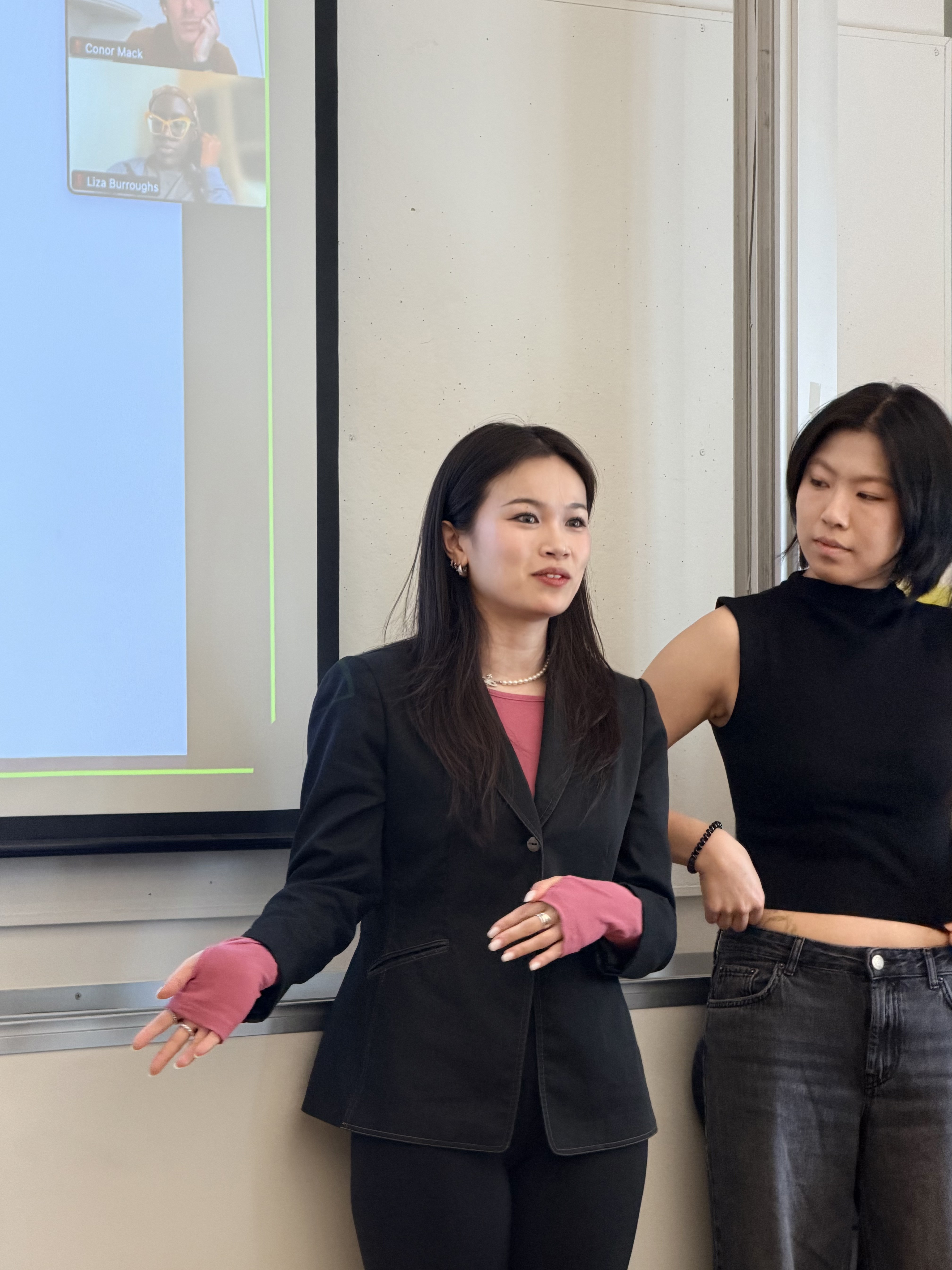 Two women stand near a screen displaying video call participants. One gestures while speaking, conveying a collaborative and focused atmosphere.
