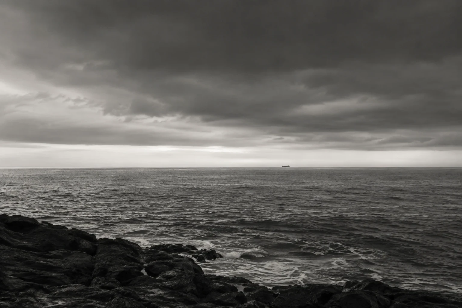 Black and white moody seascape under a broad heavy cloud bank, a tiny anonymous distant ship near horizon