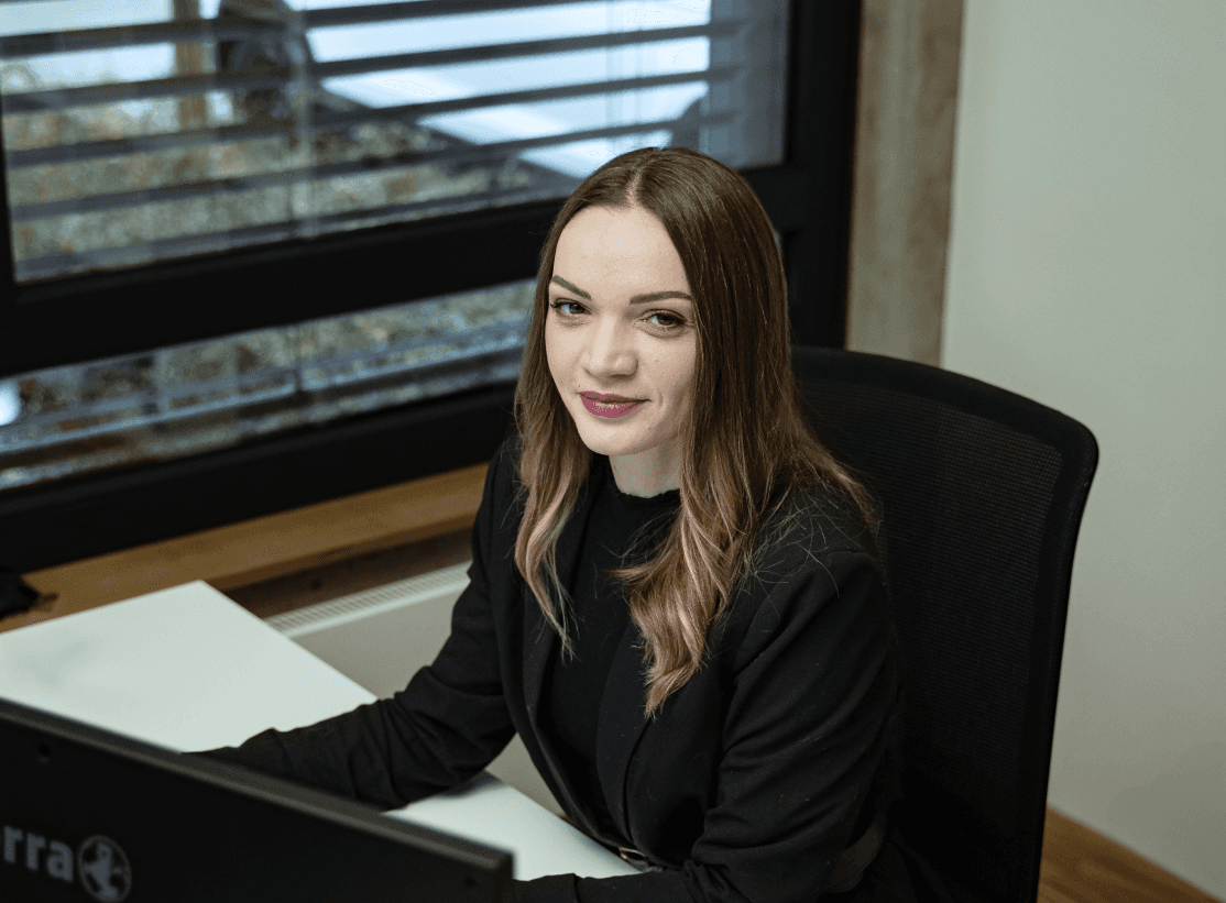 Junge Frau in schwarzem Blazer sitzt an ihrem Arbeitsplatz im Büro der kba Architekten und Ingenieure GmbH und arbeitet konzentriert am Computer; im Hintergrund großes Fenster mit Jalousien.