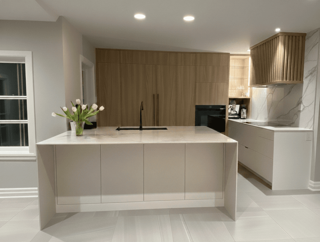 Cuisine contemporaine et minimaliste. En avant plan, un gros îlot blanc crème avec comptoir en cascade accueille un évier noir sous plan et un robinet noir, avec un beau bouquet de tulipes blanche posé sur le coin gauche. Sur le mur du fond de la cuisine, un mur complet d'armoires en finition bois clair sans poignée si trouve. Le réfrigérateur est dissimulé dans cette partie de la cuisine. Juste à côté se trouve le four encastré noir. À côté du four, légèrement en hauteur, une armoires vitrée éclairée met en valeur de la vaiselle en verre. Sur le mur de droite, la cuisine se poursuit en blanc crème avec un comptoir et un dosseret en quartz veinuré, une plaque de cuisson est sur le comptoir de quartz. Une magnifique hotte décorative  en bois si trouve, elle a un fini rainurée.