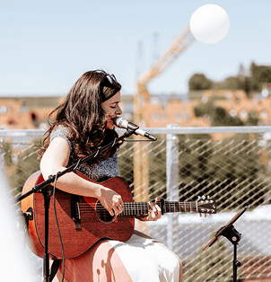 Eine Musikerin spielt konzentriert Gitarre und singt in ein Mikrofon. Sie trägt ein schimmerndes Oberteil und einen fließenden Rock. Die Bühne befindet sich im Freien, mit einem Baukran und einer urbanen Landschaft im Hintergrund. Eine weiße Dekokugel schwebt über ihr, während sie in die Musik vertieft ist. Maria Wiethaler freie Rednerin und Sängerin für Hochzeiten, Taufen, Trauerfeiern und Events aus München und Landshuter Umgebung
