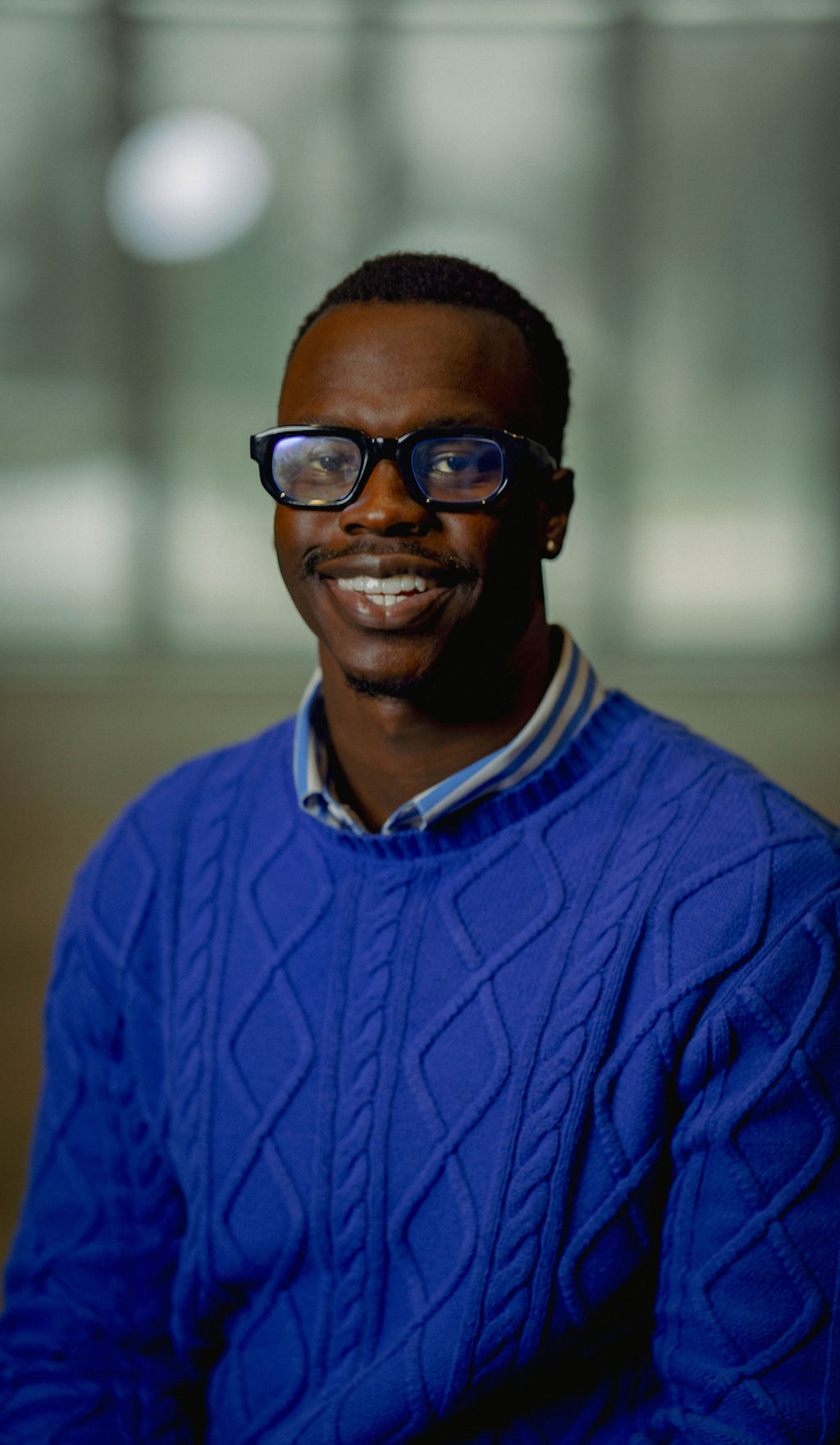 Portrait of a person with red hair, wearing a rust-colored outfit and scarf, set against a neutral gray background.