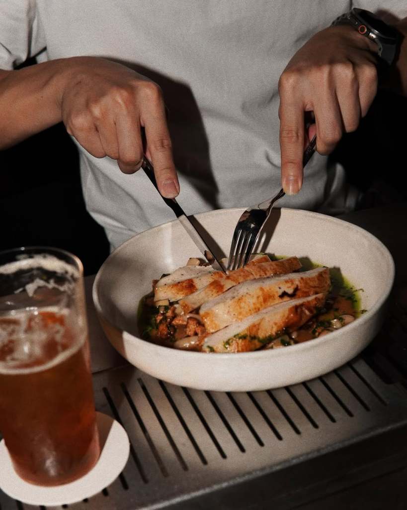 A person holding a knife and fork, preparing to eat sliced chicken on a plate, with a drink in a glass nearby.