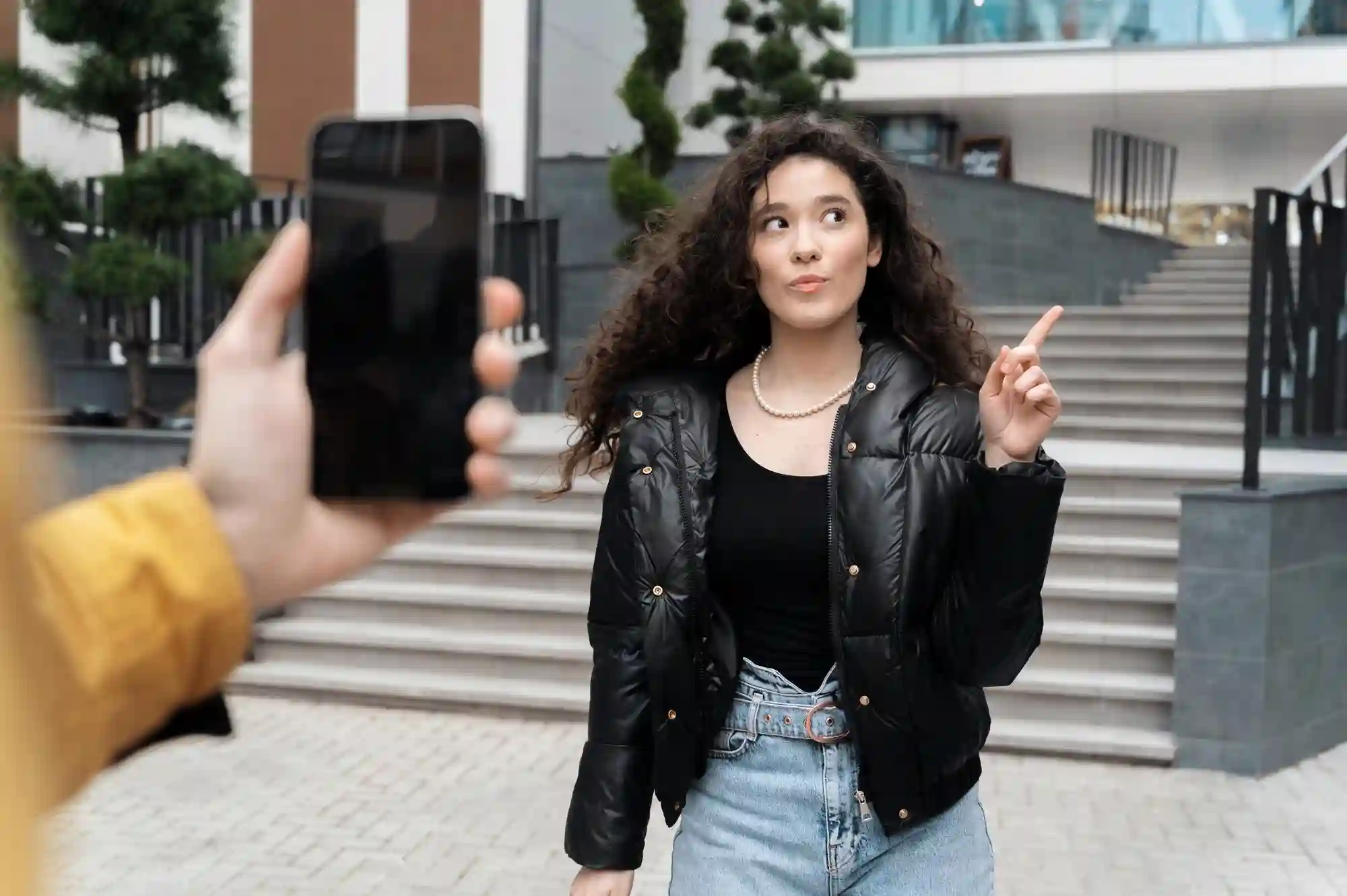 Woman posing and pointing while a person records her with a smartphone in an urban setting.